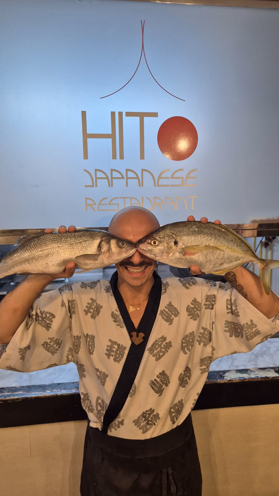 El chef de Hito sosteniendo dos pescados frescos delante del logo del restaurante