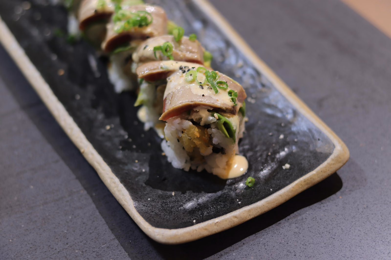 Uramaki coronado con pescado flameado y cebolleta en un plato negro en Hito