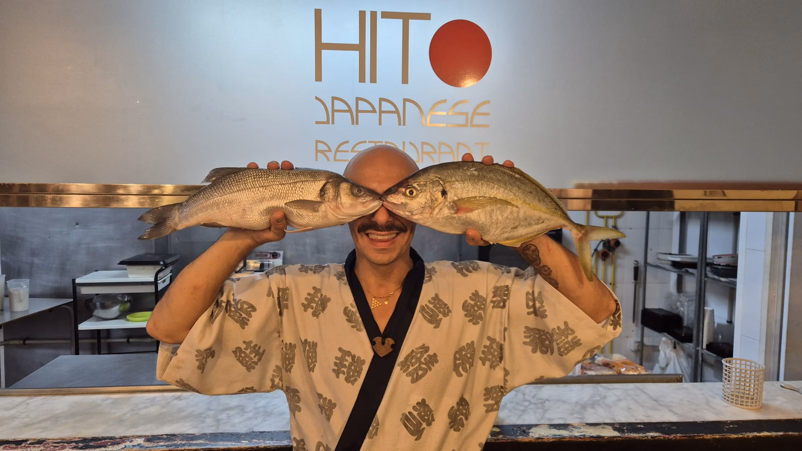 Un chef de sushi de Hito sosteniendo dos pescados frescos frente al logo del restaurante